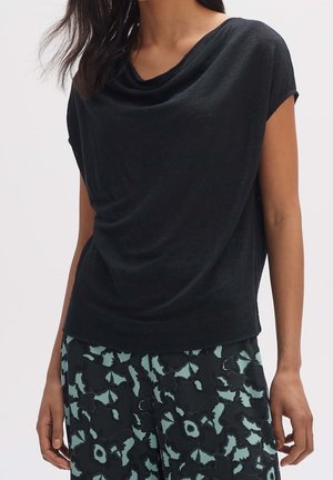 Femme portant un haut noir à manches courtes ample, associé à un pantalon noir à motifs floraux verts.