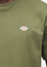 Πράσινο ελιάς μπλουζάκι από βαμβάκι, με ένα μικρό, πολύχρωμο κυκλικό λογότυπο με το κείμενο "Dickies" στην αριστερή πλευρά του στήθους.