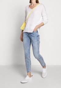 Weißer, langärmeliger V-Ausschnitt-Pullover; hellblaue Skinny-Jeans; weiße Sneakers; gelbe Umhängetasche. Lässiges Outfit mit unterschiedlichen Texturen.