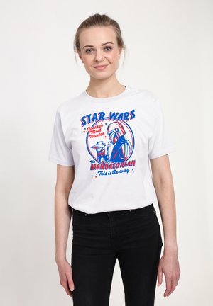 Junge Frau trägt ein weißes Star Wars T-Shirt mit Mandalorian und Grogu sowie dem Text "Galaxy's Most Wanted" und "This is the way."