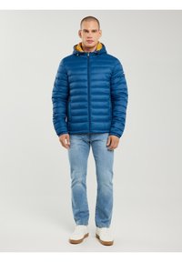 Férfi, kék steppelt dzsekivel, sárga kapucnis béléssel, világoskék farmerrel és fehér tornacipővel, sima háttér előtt áll.