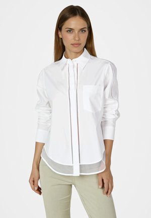 Frau trägt ein weißes Hemd mit Knöpfen und Brusttasche sowie sichtbaren Nahtdetails, kombiniert mit hellbeigen Hosen, stehend vor einem schlichten Hintergrund.