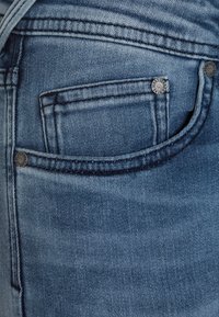 Jeans de denim en azul deslavado, con un pequeño bolsillo frontal, remaches de bronce y costuras en contraste, mostrando una textura suave.