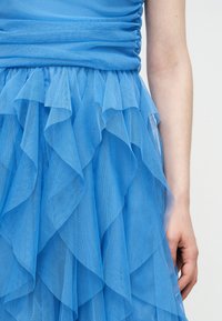 Detail modrej viacvrstvovej tylovej sukne so zošitým pásom, zobrazený na dolnej časti trupu a pravom ramene osoby na jednotnom pozadí.