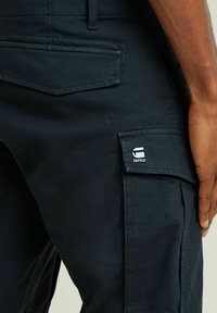 Ciemnoniebieskie spodnie cargo z teksturowanej tkaniny, z wieloma kieszeniami i widoczną łatą z logo w okolicy talii.