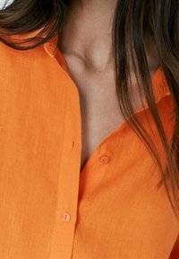 Frau trägt ein oranges Hemd mit Knöpfen, lange braune Haare bedecken teilweise den Kragen- und Schulterbereich.