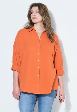 Frau mit langem braunem Haar trägt ein lockeres orangefarbenes Hemd mit aufgekrempelten Ärmeln und blaue Jeans, steht vor weißem Hintergrund.