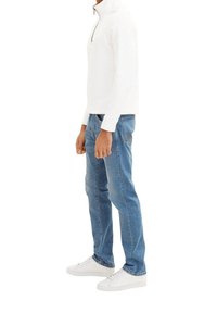 Camisa blanca de manga larga con cremallera, jeans azules y zapatillas blancas. La camisa tiene un cuello alto; los jeans son ligeramente ajustados con costuras visibles.
