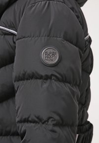 Detalle de una chaqueta acolchada negra con un parche redondo y texturizado con el logo "SUPERDRY" y textura de tela que muestra un acabado brillante y un patrón cosido.