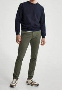 Uomo che indossa un felpa blu navy, pantaloni verde oliva e scarpe beige e marroni, in piedi contro uno sfondo chiaro e semplice.