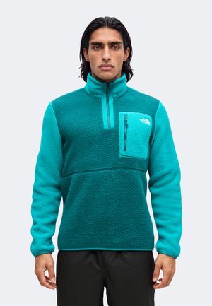 Mand iført en fleecetrøje i teal og turkis med lynlåslomme på brystet, stående front mod kameraet mod en ensfarvet lys baggrund.
