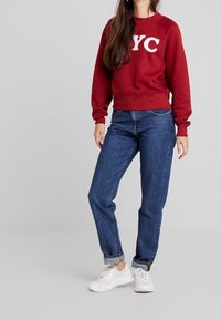 Burgundy sweatshirt with white "NYC" text, paired with blue jeans and white sneakers. The sweatshirt has a ribbed neckline and cuffs.