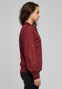 Blouson aviateur bordeaux avec une texture lisse et brillante. Doté d'un col côtelé, de poignets et de poches latérales, présenté en profil latéral.