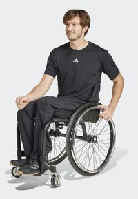 Černé sportovní tričko s bílým logem, černé kalhoty a černé tenisky; sedící na invalidním vozíku s kovovými koly a gumovými pneumatikami.