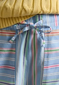 Pull jaune tricoté porté sur un short en tissu bleu avec des rayures pastels verticales et horizontales, noué par un nœud à la taille.