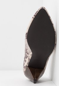 Chaussure à talon haut pour femme à bout pointu avec un motif peau de serpent et une semelle sombre, positionnée verticalement contre un fond blanc en angle.