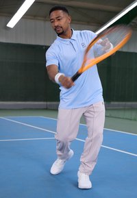 Lyseblå polo skjorte, beige treningbukser og hvite joggesko. Spilleren holder et oransje tennisracket og er i en dynamisk svingebevegelse på en blå bane.