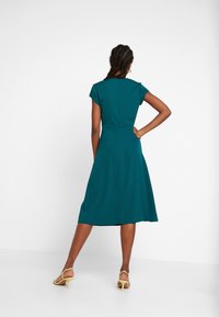 Teal-huulinen mekko, jossa on lyhyet hihat, istuva muotoilu ja leveä polvipituinen hame. Tasainen kangas, jossa vetoketju takana. Keltaiset korkokengät.