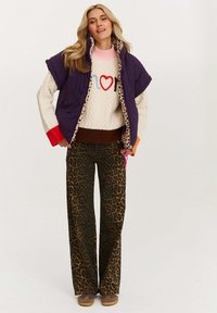 Fioletowa kamizelka pikowana na kremowym swetrze w wzory z motywem serca, połączona z szerokimi spodniami w panterkę i brązowym obuwiem.