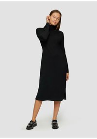 Robe noire à manches longues avec col montant, présentant une coupe droite et une fente latérale. Portée avec des mocassins noirs, texture lisse et design minimal.