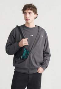 Γκρι φούτερ με φερμουάρ και απαλή υφή, με δύο εμπρόσθιες τσέπες και μικρό λογότυπο. Συνδυάζεται με σκούρο πουκάμισο και τσάντα ώμου σε τιρκουάζ χρώμα.