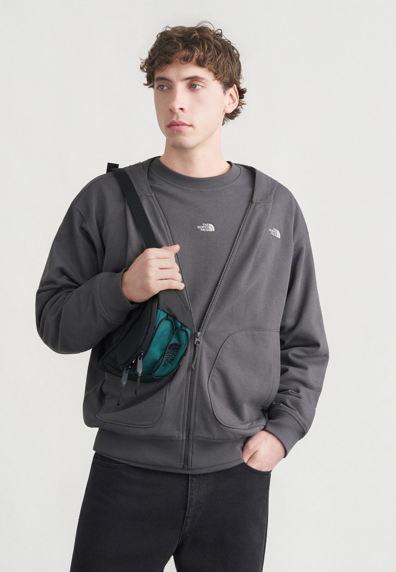 Γκρι φούτερ με φερμουάρ και απαλή υφή, με δύο εμπρόσθιες τσέπες και μικρό λογότυπο. Συνδυάζεται με σκούρο πουκάμισο και τσάντα ώμου σε τιρκουάζ χρώμα.