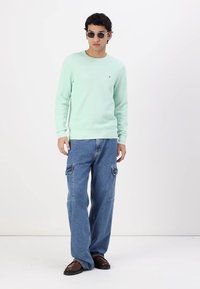 Pull mint à col rib végétal, associé à un jean cargo bleu clair avec poches latérales et des mocassins marron. Lunettes de soleil portées.