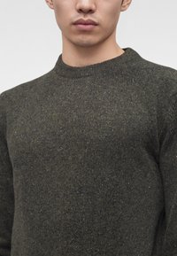 Gros plan sur une personne portant un pull en tricot vert foncé moucheté avec un petit logo brodé sur la poitrine.