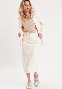 Jupe midi crème en coton, assortie à un top beige à col en V et une légère veste. La tenue présente des textures discrètes et des teintes neutres.