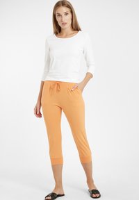 Haut blanc à manches longues associé à un pantalon court orange avec une taille élastique et un cordon de serrage. Des sandales plates noires complètent la tenue.