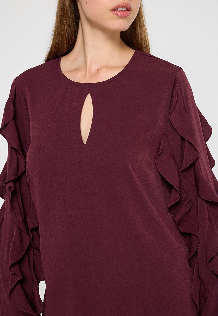 Bordeaux blouse met een ronde hals, sleutelkastje-detail en gerimpelde mouwen. Gladde stof met een lichtvallende pasvorm.