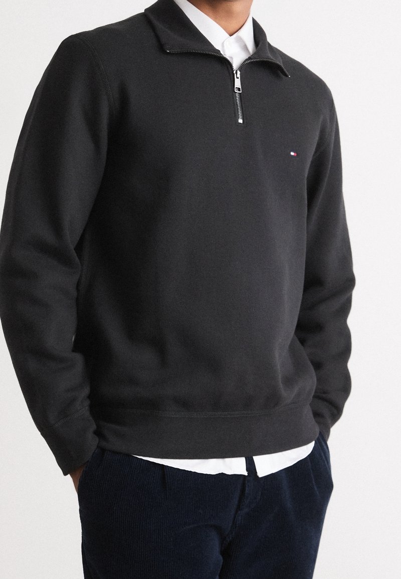 Homme portant un pull noir à fermeture éclair quart de zip sur une chemise blanche avec un pantalon bleu marine, mains dans les poches, fond clair uni.