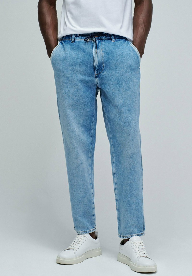 Pantalones de mezclilla azul claro con un corte relajado, que cuentan con bolsillos frontales y una cintura elástica con cordón, combinados con zapatillas blancas.