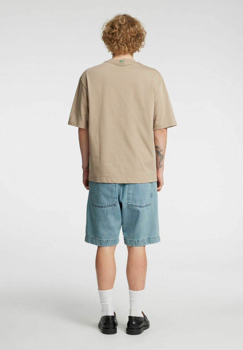 Beige bomulls-T-skjorte med løs passform og korte ermer, kombinert med lyseblå denimshorts med to baklommer. Svarte sko og hvite sokker.