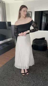 Haut noir en dentelle à épaules dénudées, jupe longue plissée argentée, talons noirs à lanières, debout sur un tapis gris texturé dans un intérieur moderne.