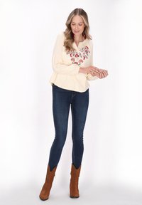 Blusa color crema con design floreale ricamato, maniche lunghe e vestibilità rilassata, abbinata a jeans skinny scuri e stivaletti alla caviglia in suede marrone.