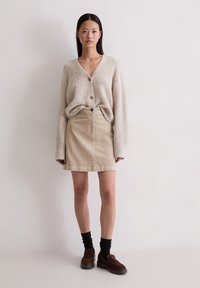 Beżowy sweter warkoczowy z teksturowanej tkaniny i dużymi guzikami, zestawiony z beżową spódnicą dżinsową, czarnymi skarpetkami i brązowymi butami wsuwanymi.