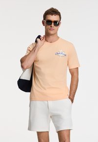 Lyserød bomulds t-shirt med "Club Locale" tryk i blå, parret med hvide shorts. Model holder en sort og hvid taske.