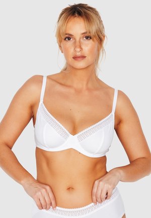Femme à la peau claire et aux cheveux blonds portant un soutien-gorge blanc bordé de dentelle et une lingerie assortie, debout devant un fond uni.