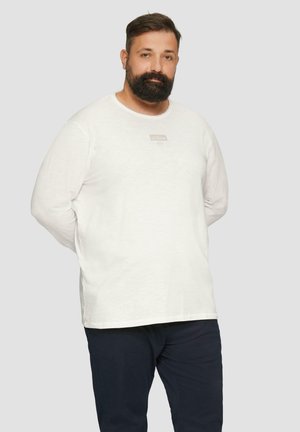 Witte longsleeve t-shirt met een ronde halslijn en een klein beige logo op de borst, gedragen met donkere broek tegen een neutrale achtergrond.