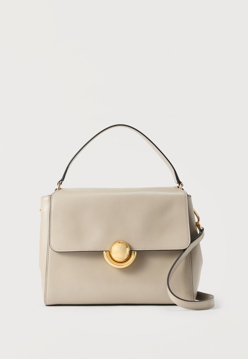 Beige Ledertasche mit einem oberen Griff, Klappenverschluss, goldfarbenem Rundverschluss und abnehmbarem Schulterriemen vor weißem Hintergrund.