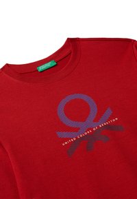 T-shirt di cotone rossa con scollatura rotonda. Presenta un design grafico blu e la scritta "UNITED COLORS OF BENETTON" sul davanti.
