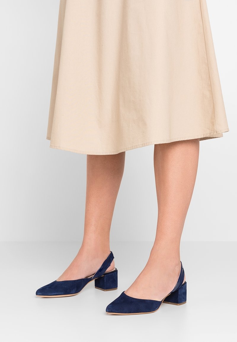 Jambes portant une jupe midi beige et des chaussures à talons blocs en daim bleu marine avec brides arrière, sur un sol blanc lisse.