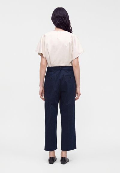 Femme aux longs cheveux foncés portant un chemisier blanc à manches courtes, un pantalon bleu marine taille haute et des chaussures noires à enfiler, debout de dos.