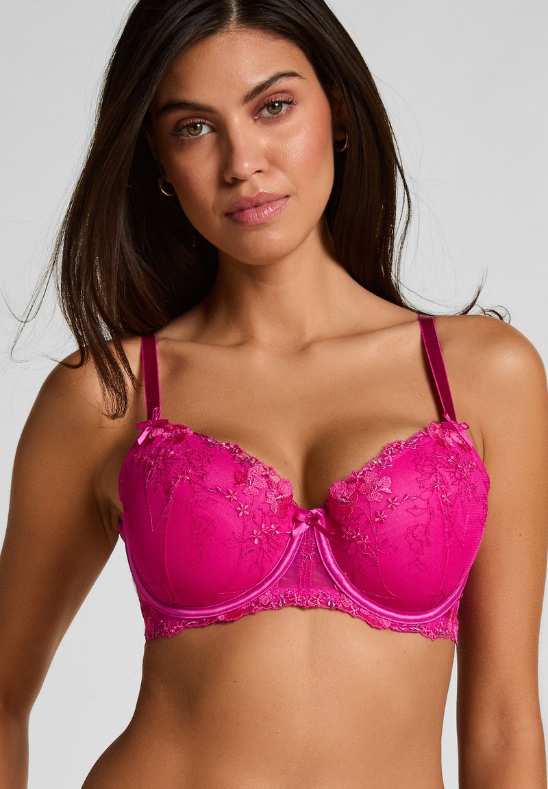 Femme portant un soutien-gorge en dentelle rose vif avec broderie florale, bretelles ajustables et petit nœud central, posant devant un fond uni.