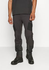 Svarte cargo-bukser med kontrasterende nylon-detaljer, mesh-paneler på nedre ben og oransje glidelåsdetaljer. Brukt med svarte snøreb boots.