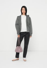 Pilkas užtrauktukas džemperis ant baltos marškinėlių, juodi džinsai, šviesiai rožinės basutės ir levandų spalvos raištelių krepšys, stovintis prieš baltą foną.