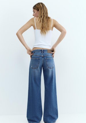 Žena s dlhými vlasmi si upravuje voľné modré rifle, oblečená v bielom tielku bez rukávov, stojaca pred jednoduchým bielym pozadím.