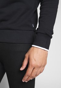 Felpa nera con polsini e orlo a costine, con un accento bianco lungo il polsini. Il tessuto appare morbido con una texture liscia.