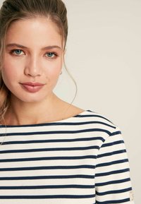Gestreiftes Boot-Hals-Top mit navyblauen und weißen horizontalen Streifen. Ripptextur und ein kleines Logo-Patch am Ärmel.
